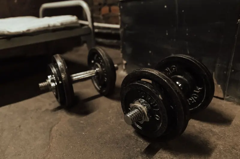 A pair of dumbbells on a concrete floor beside a metal bed in a dark, indoor setting