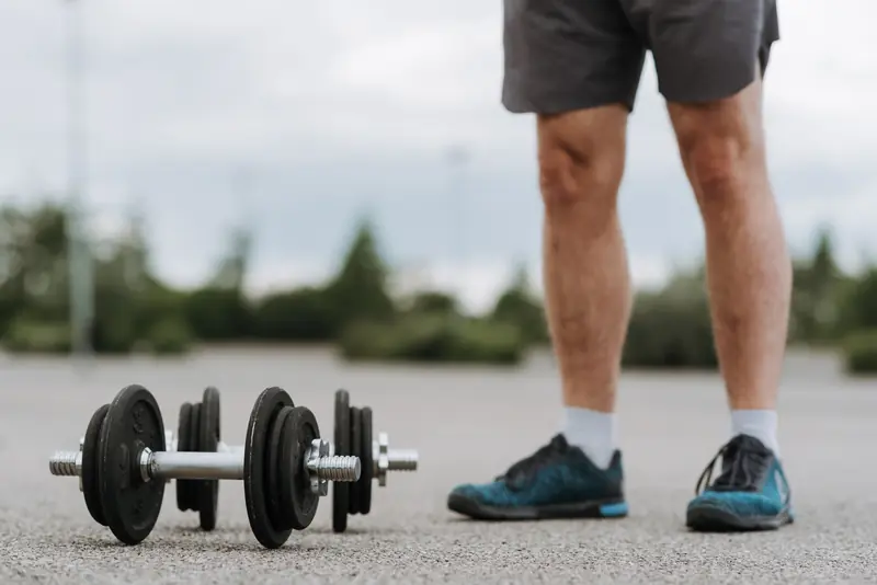 Crop anonymous male in sportswear standing near heavy metal dumbbells on asphalt road