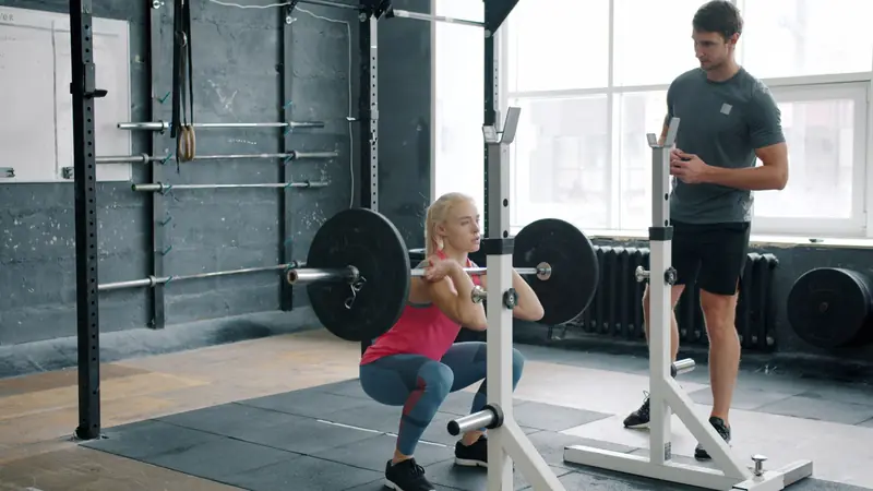 A woman performs a front squat with a barbell, guided by a trainer in a gym