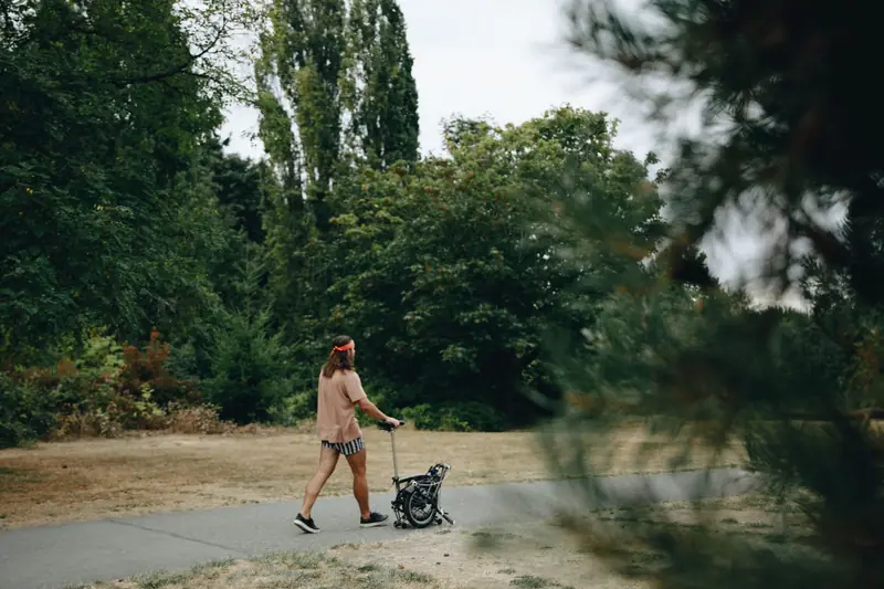 Man pushing folding bike on park path surrounded by green trees in summer, enjoying a leisurely walk