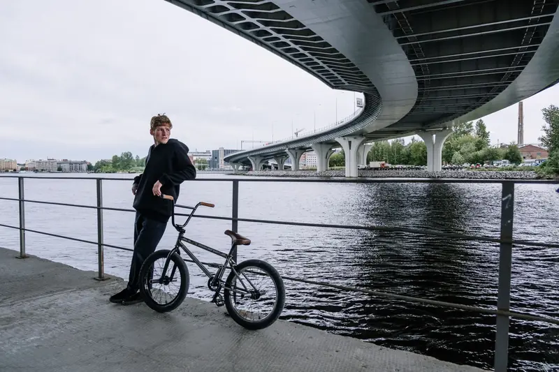 A man leaning on a railing with a BMX bike under a city bridge by the river